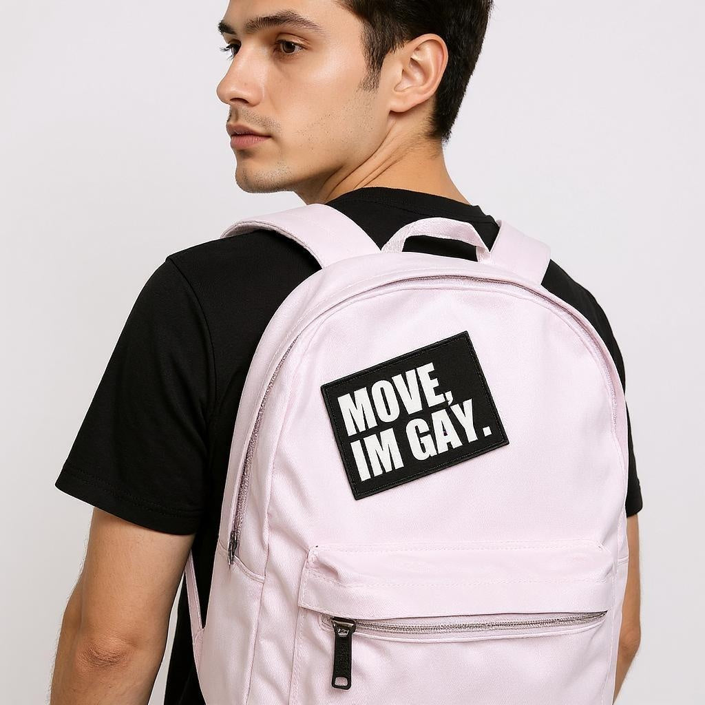 Young man wearing a light pink backpack with a bold black-and-white patch that reads “MOVE, I’M GAY.” The backpack is worn over a black t-shirt, and the overall look is casual, confident, and queer-affirming.