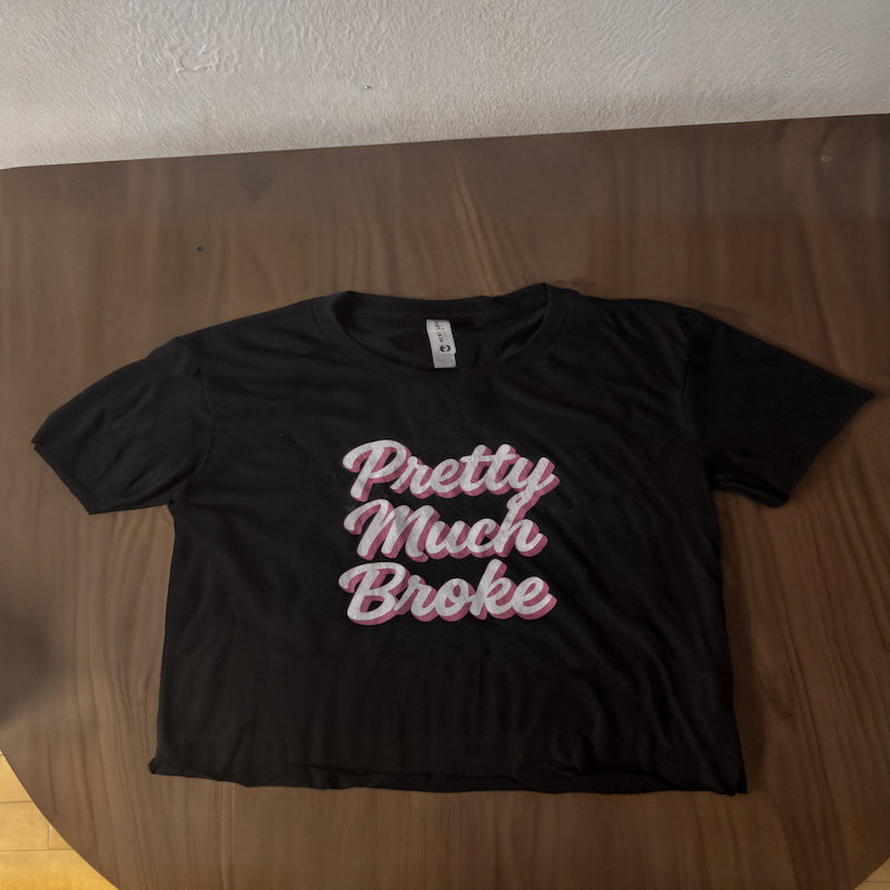 A black crop top with the text "Pretty Much Broke" on a brown wooden table.