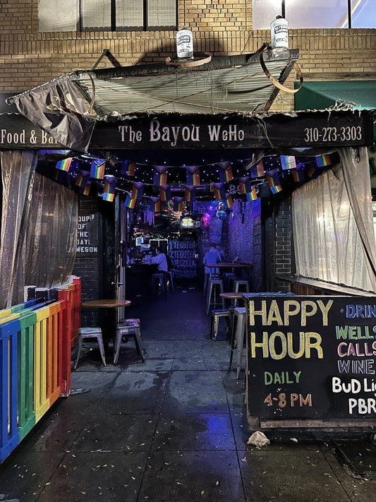 The Bayou WeHo bar entrance on Santa Monica Blvd, West Hollywood — rainbow decor, Pride flags, and happy hour sign (4–8 PM) promoting cheap drinks and Cajun seafood. Popular affordable gay bar and best happy hour spot in Los Angeles.