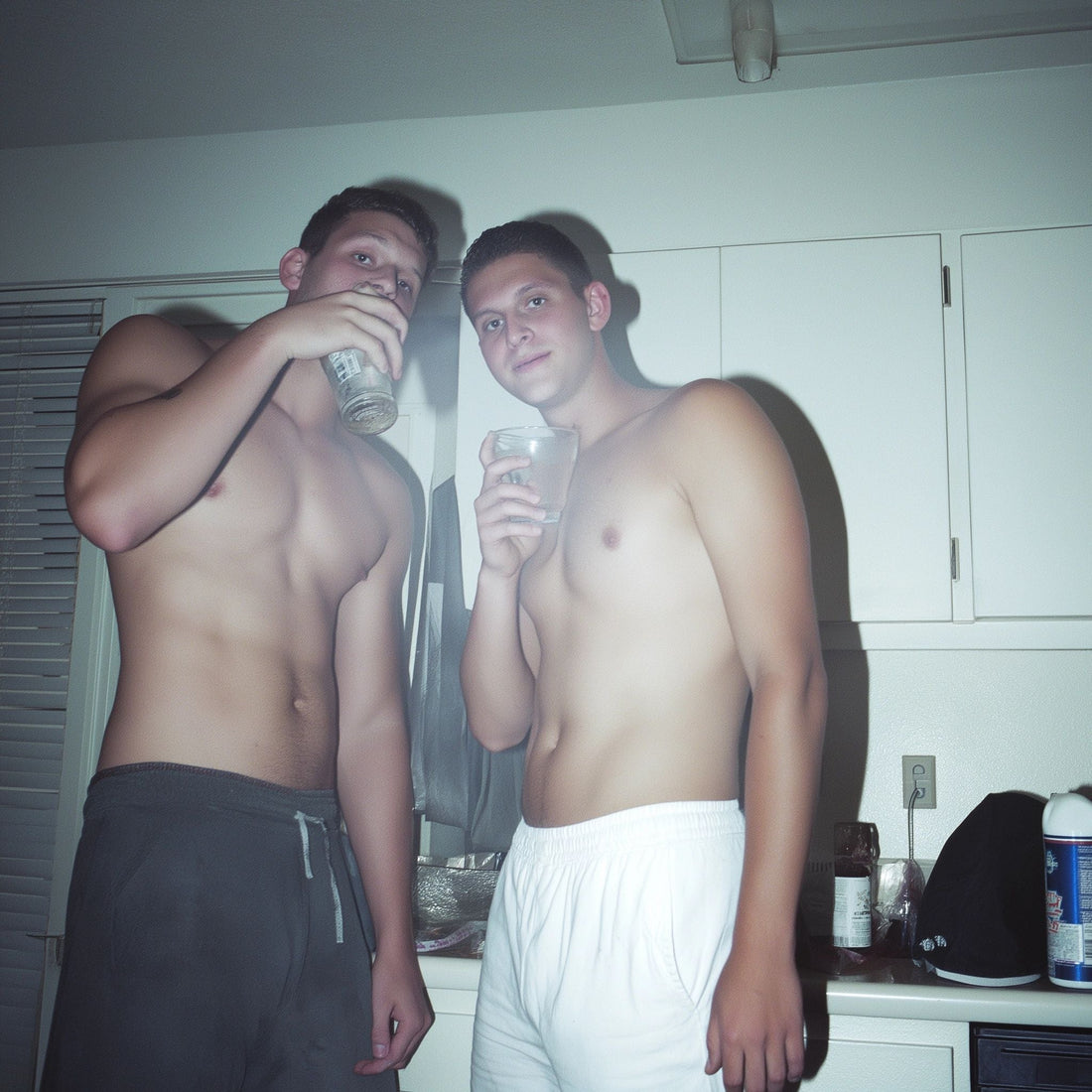 Two shirtless young men hanging out in a kitchen, casually sipping drinks, showing off confident queer energy and relaxed masculine style—capturing the vibe that inspires The Bayou WeHo’s LGBTQ+ apparel and gay fashion aesthetic.