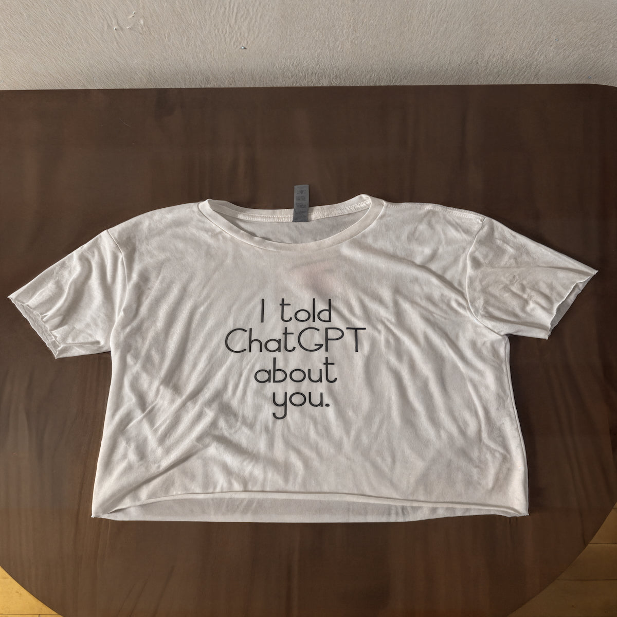 White t-shirt with text "I told ChatGPT about you" on a wooden surface