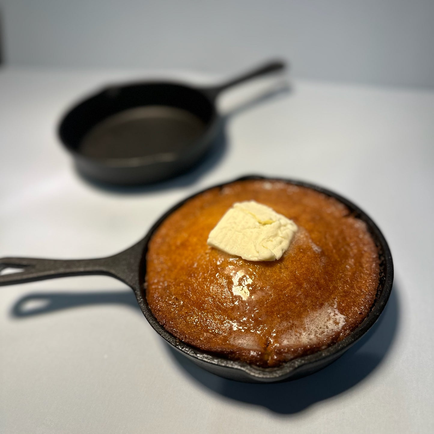 A cast iron skillet with a honey butter cornbread muffin on it, with a pat of butter on top. Another empty skillet is visible in the background.