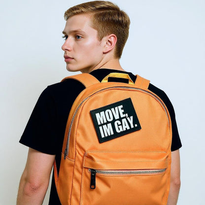 Person wearing an orange backpack with a 'MOVE, IM GAY.' patch on a plain background
