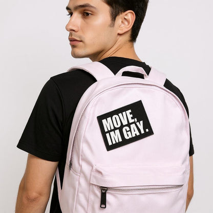 Young man wearing a light pink backpack with a bold black-and-white patch that reads “MOVE, I’M GAY.” The backpack is worn over a black t-shirt, and the overall look is casual, confident, and queer-affirming.