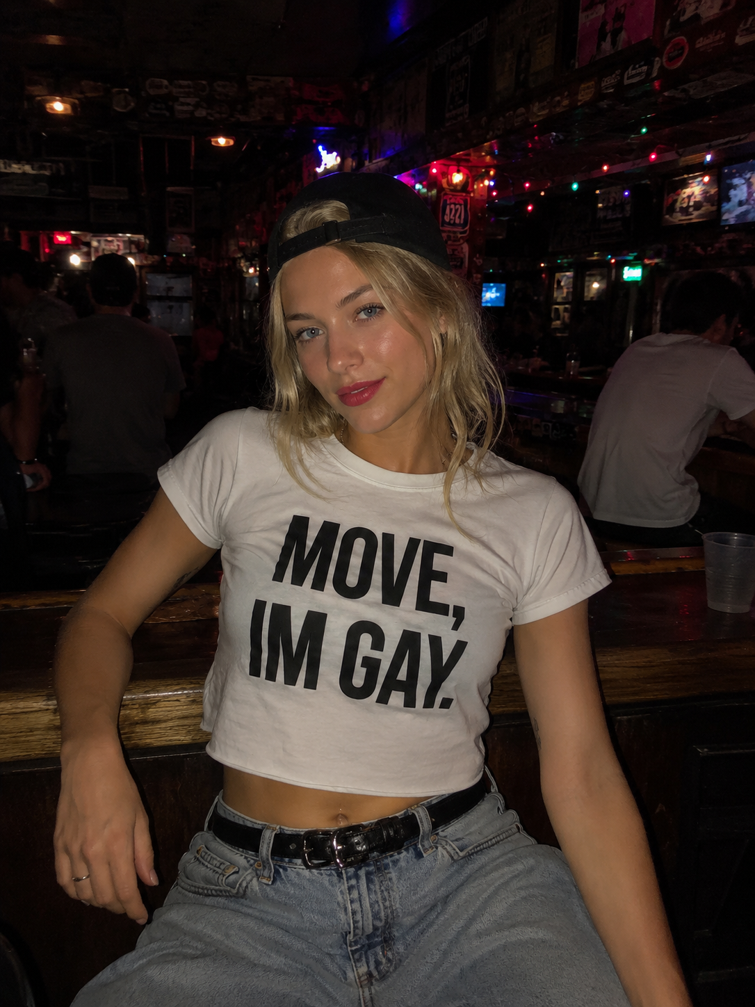 Person wearing a t-shirt with 'MOVE, I'M GAY.' text in a bar setting