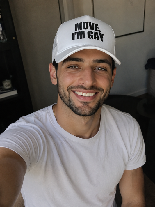 A smiling man wearing a white trucker-style cap with the text 'MOVE, I'M GAY' in black letters, paired with a white t-shirt in an indoor setting.