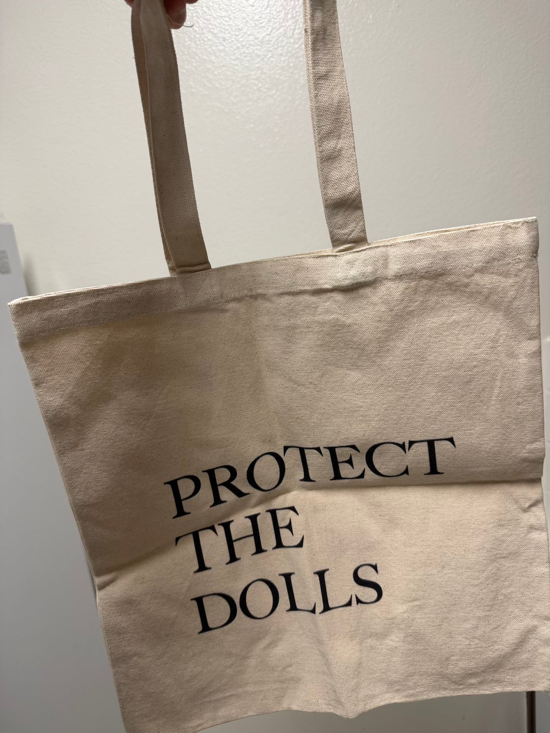 Beige tote bag with 'PROTECT THE DOLLS' text on a plain background
