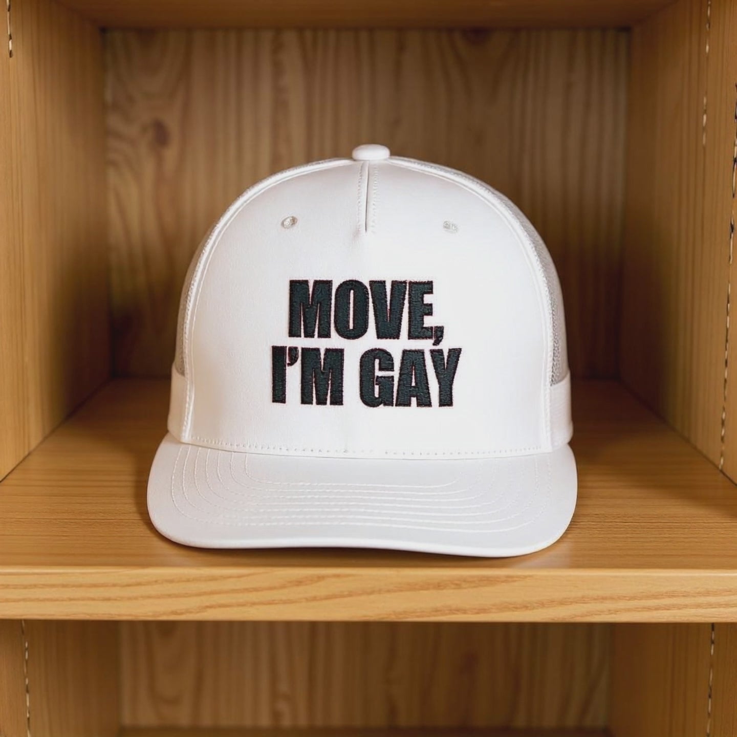 A white trucker-style hat with bold black embroidery that reads “MOVE, I’M GAY” spinning on a wooden shelf, representing queer pride apparel, bold LGBTQ+ fashion, and campy streetwear accessories.