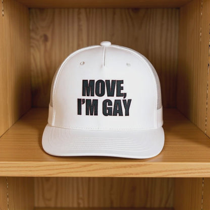 A white trucker-style hat with bold black embroidery that reads “MOVE, I’M GAY” spinning on a wooden shelf, representing queer pride apparel, bold LGBTQ+ fashion, and campy streetwear accessories.