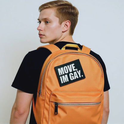 A person wearing a bold "MOVE, I'M GAY" patch on a vibrant orange backpack, showcasing unapologetic queer pride and playful LGBTQ+ fashion.
