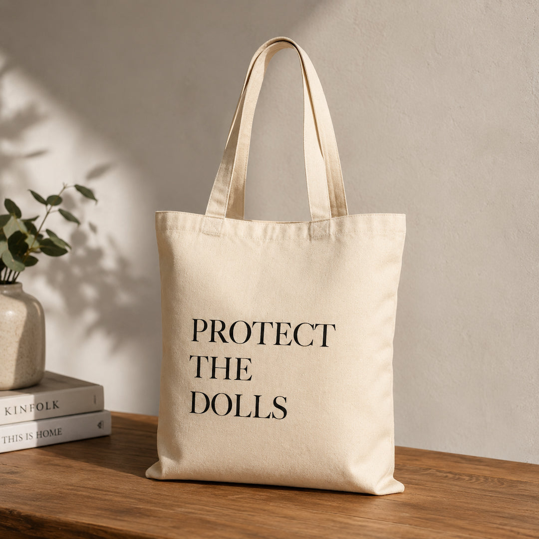 Beige tote bag with 'PROTECT THE DOLLS' text on a wooden surface with books and a plant.