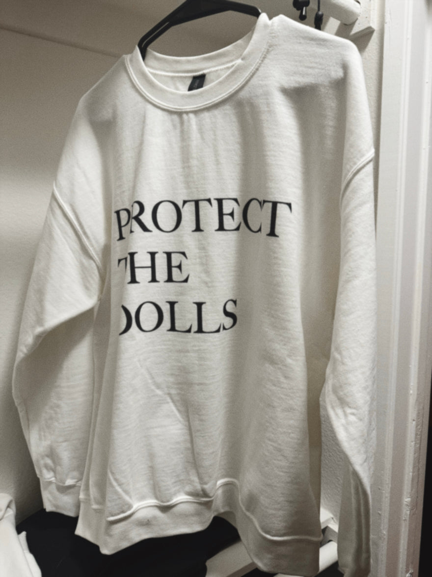 White sweatshirt with 'PROTECT THE DOLLS' text on a hanger against a white wall.